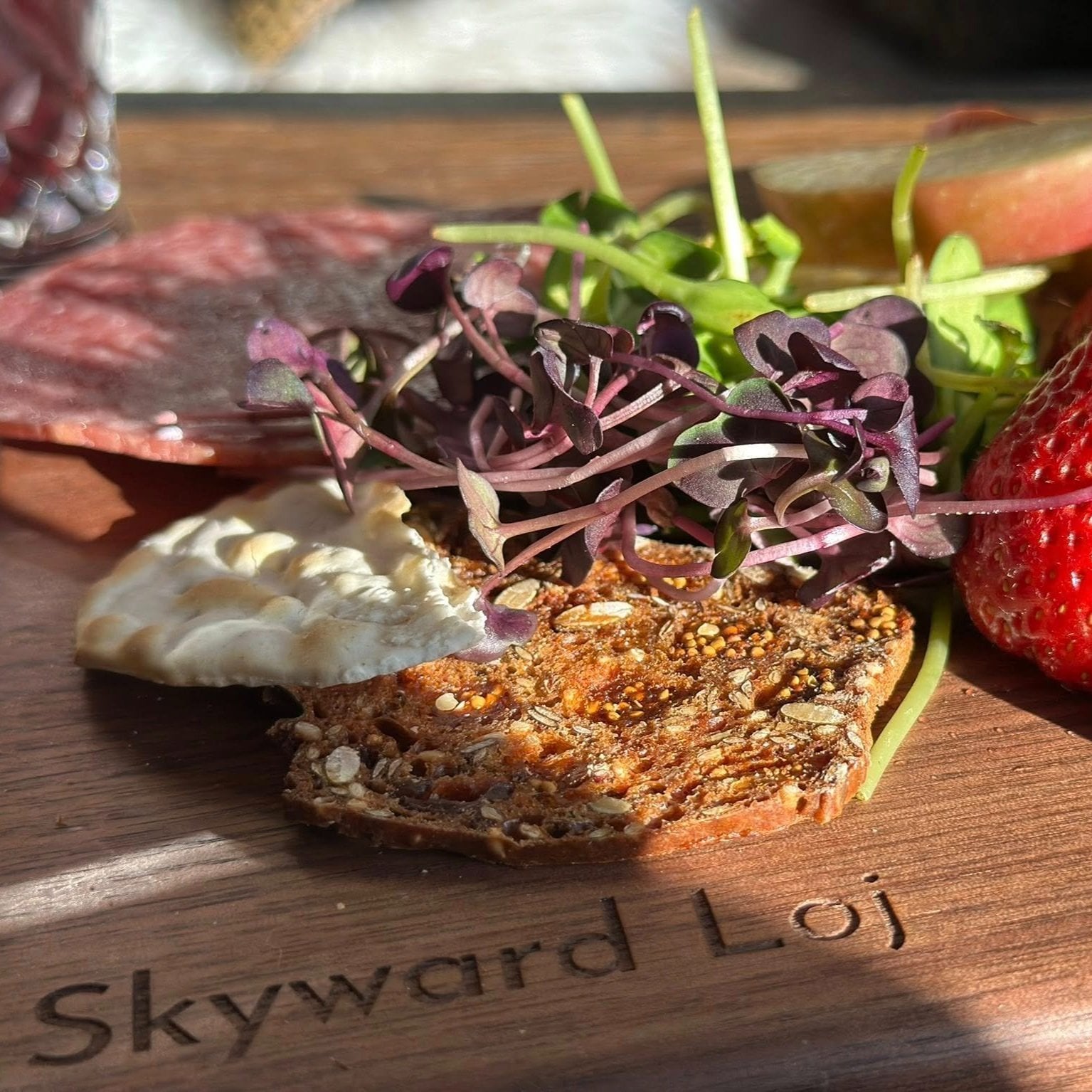 A close-up of a wooden platter featuring a variety of gourmet foods, including a slice of crispbread topped with soft cheese, fresh microgreens, strawberries, and cured meats, highlighting a vibrant and appetizing presentation.